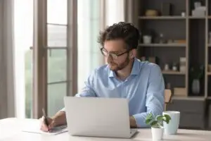 Credentialed young man sitting at table in front of computer, writing on paper